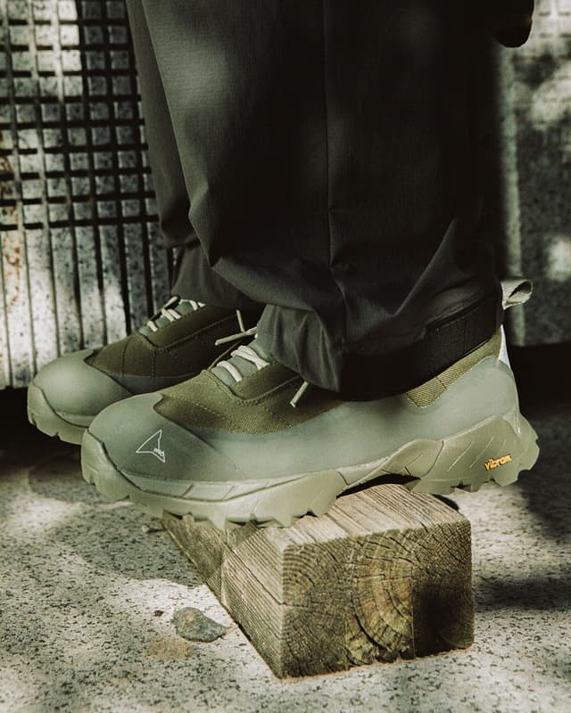 A person's feet in olive green, rugged shoes with a white logo, one foot resting on a wooden block.