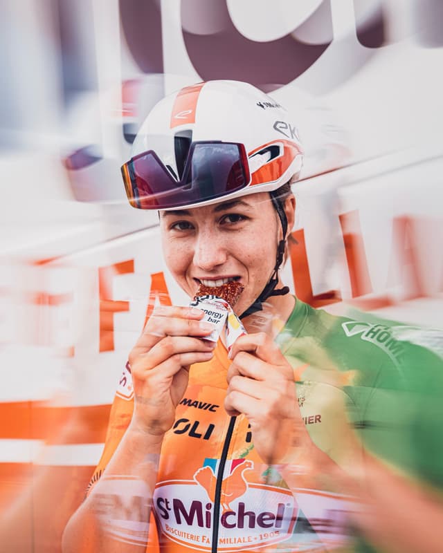 A smiling female cyclist in a helmet and orange and green team jersey eats an energy bar.