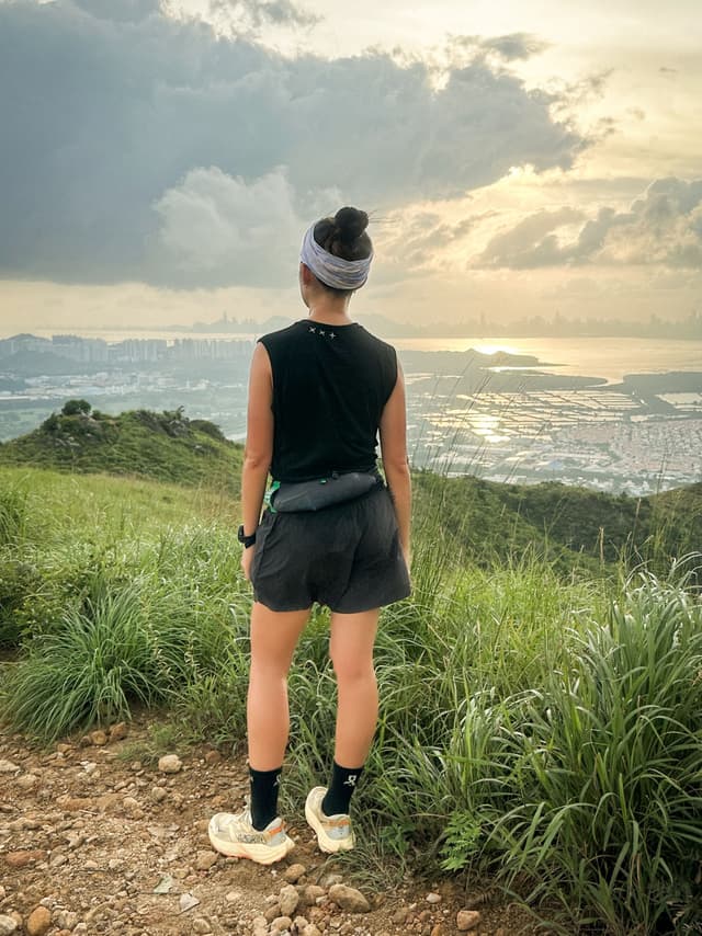 Rear view of a person on a mountain trail looking at a distant city and bay under a cloudy sunset sky.