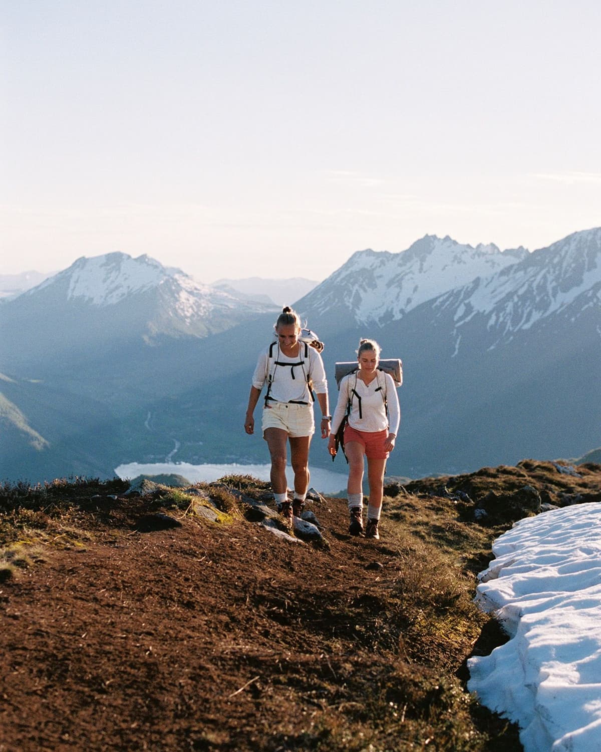 Two backpackers on a mountain trail, with snow-capped peaks and a lake below.