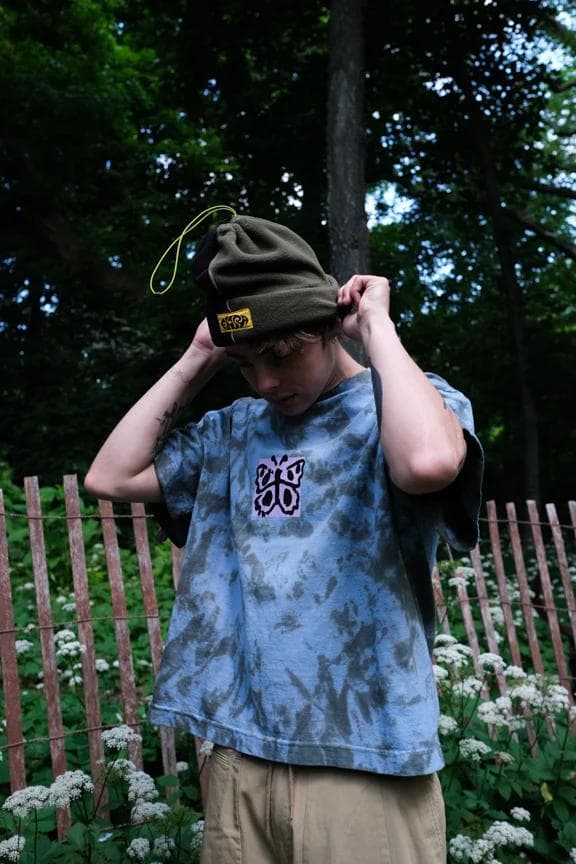 A person adjusting an olive green beanie, wearing a blue tie-dye shirt with a butterfly graphic, outdoors amidst trees and a wooden fence.