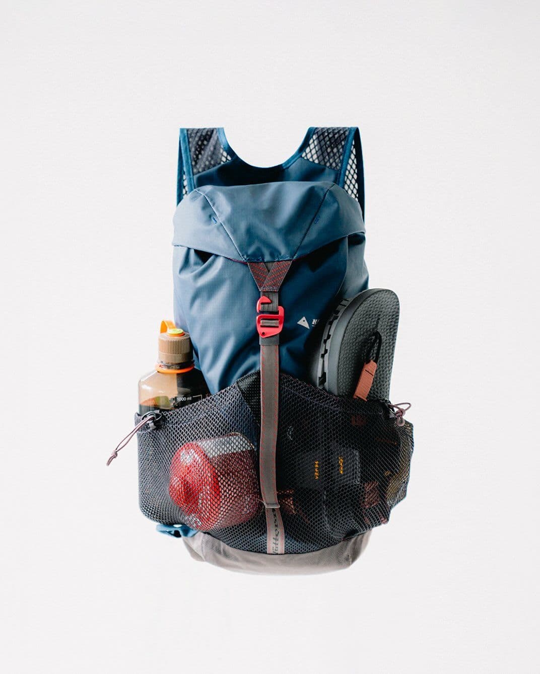 A blue trail running vest with a water bottle, red gear sack, and sandals in its mesh pockets.