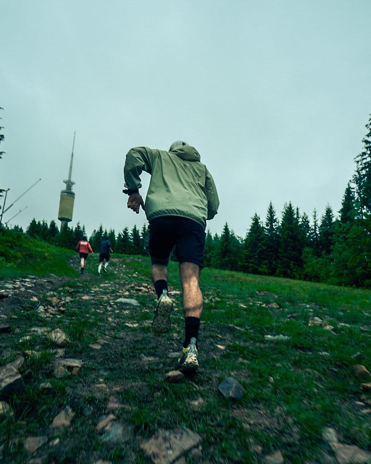 Rear view of a person trail running uphill on a rocky path with other runners, a forest, and a tower.