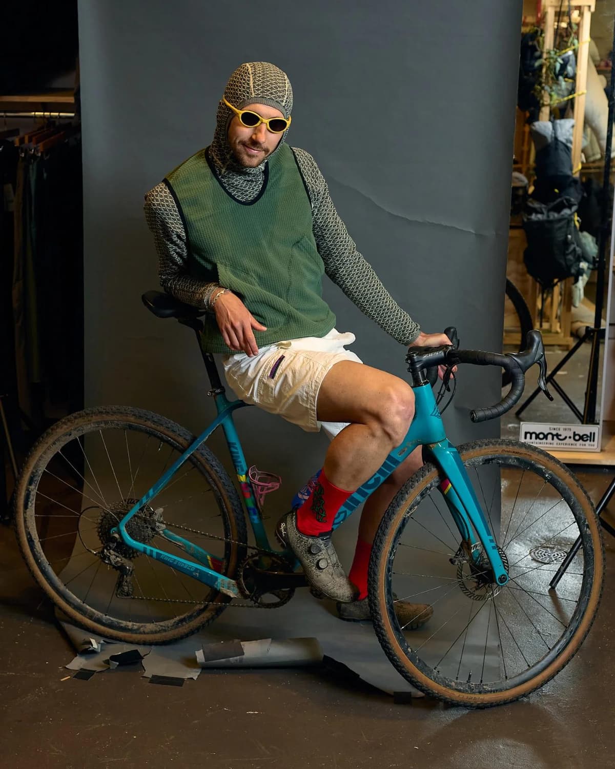 A man in a patterned balaclava, long-sleeve shirt, green vest, white shorts, and yellow sunglasses sits on a dirty teal gravel bike.