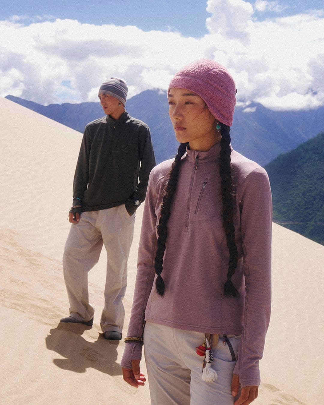 A woman in a pink beanie and braids stands on a sand dune, with a man behind her and mountains under a cloudy sky.