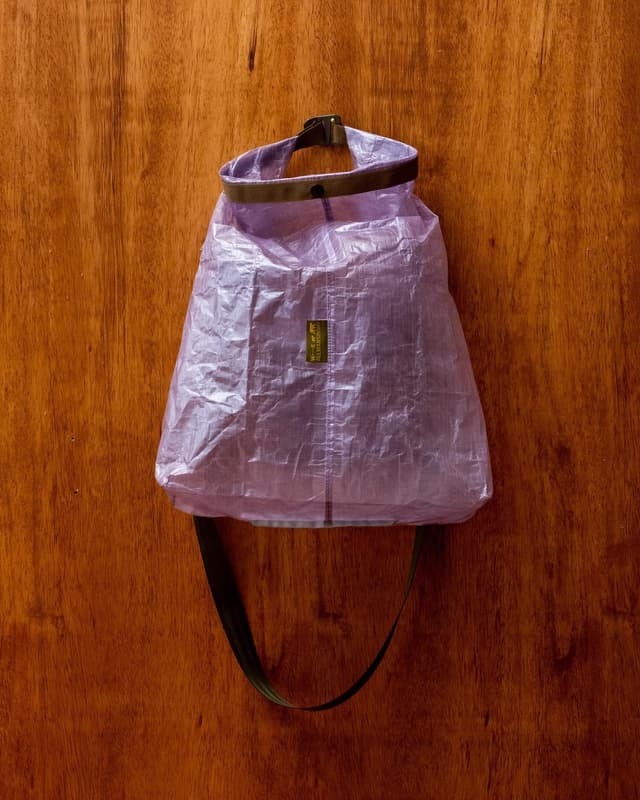 A translucent lavender roll-top dry bag with a dark strap, hanging against a wooden wall.