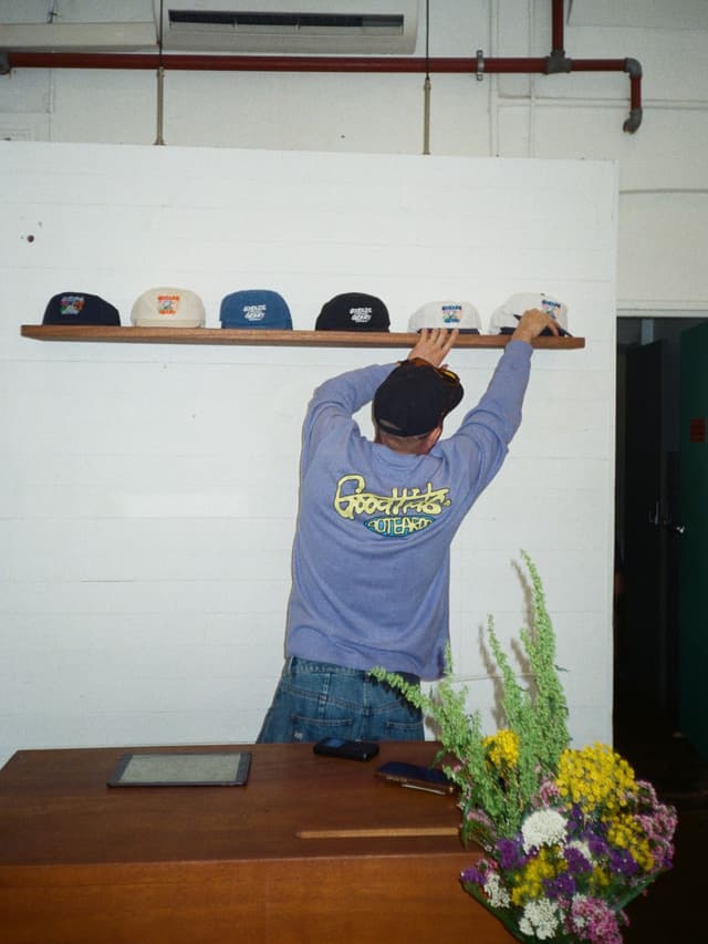 A person from behind arranges baseball caps on a shelf above a wooden counter with a colorful flower bouquet.