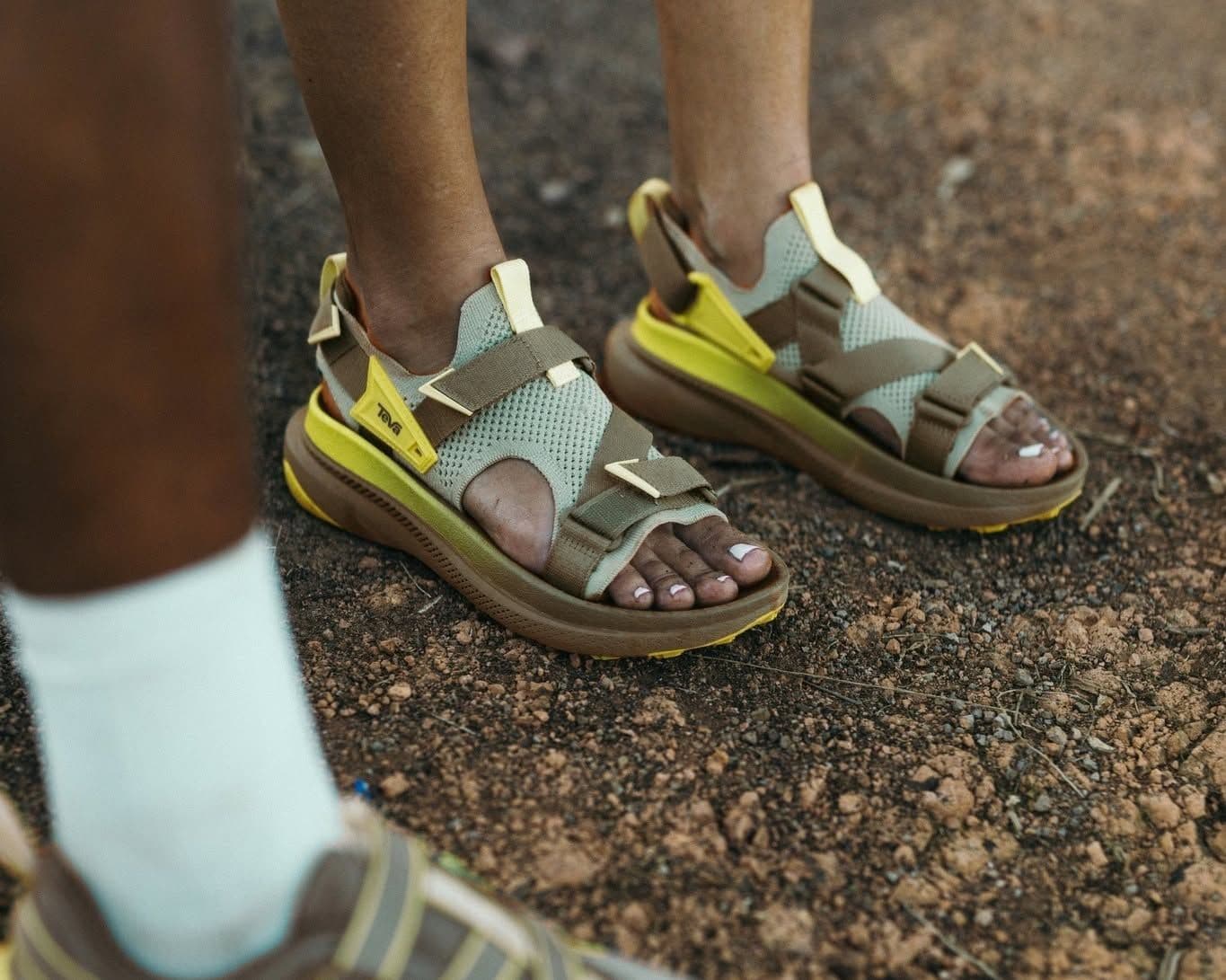 A person's feet in khaki and yellow Teva sandals on a dirt path.