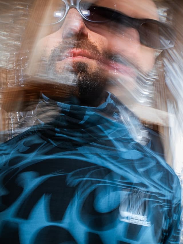 Motion-blurred man wearing sunglasses and a dark turtleneck with glowing blue patterns.