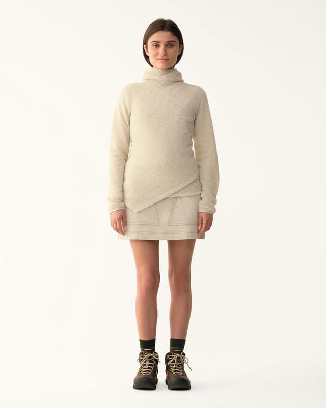 A woman in a cream fuzzy wrap-style turtleneck sweater, light mini skirt, and brown hiking boots stands on a white background.