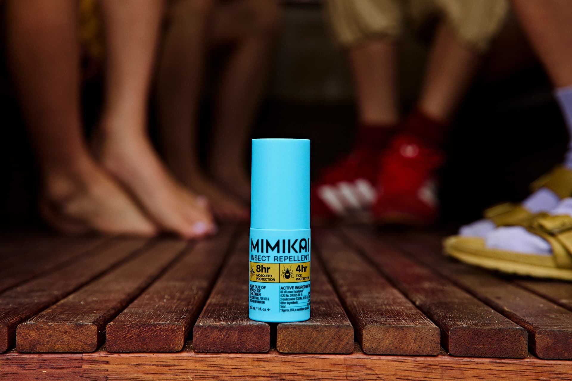 Blue Mimika insect repellent bottle on a wooden deck, with blurred feet in the background.