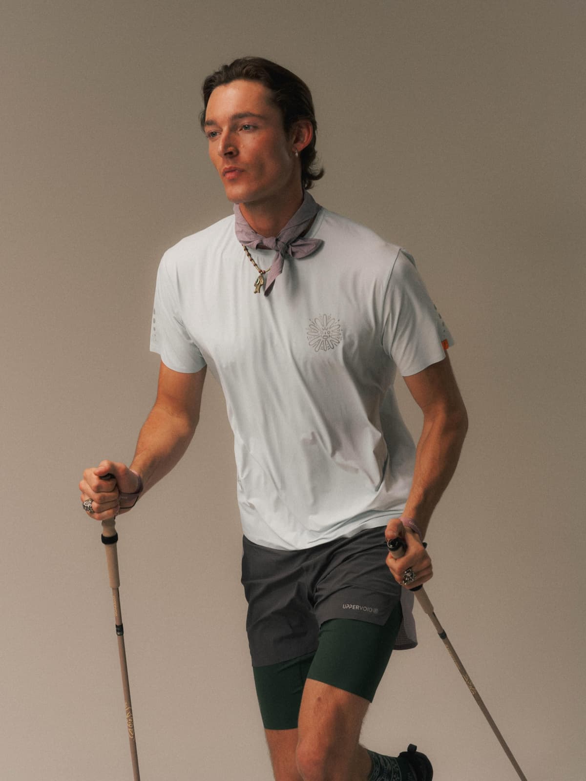 A man in a light t-shirt, bandana, and layered shorts holds two trekking poles.