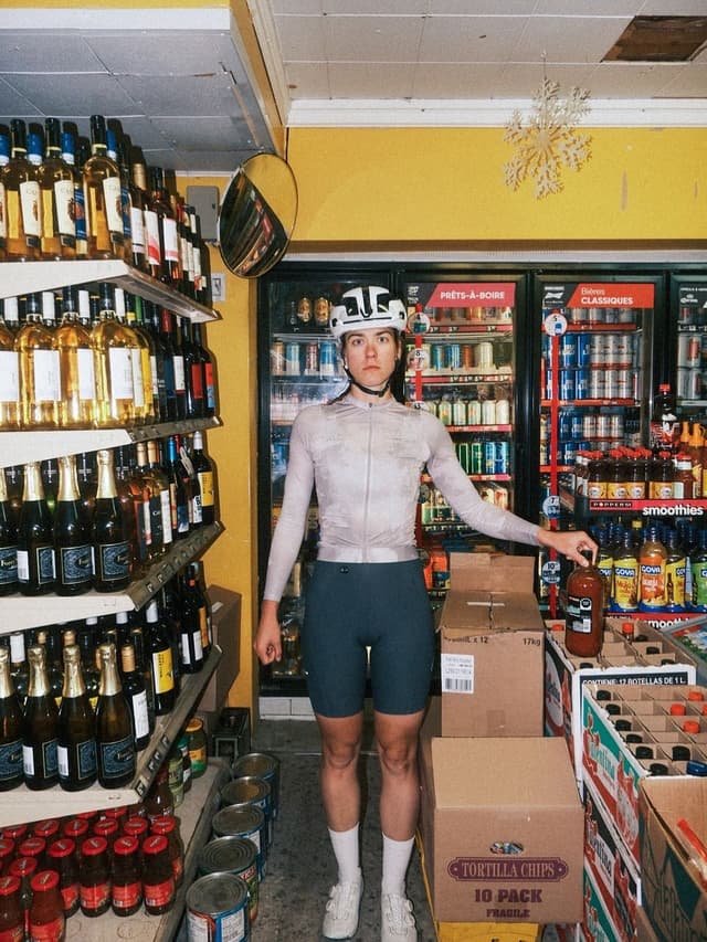 A cyclist in a helmet and full gear stands in a convenience store aisle, holding a bottle.