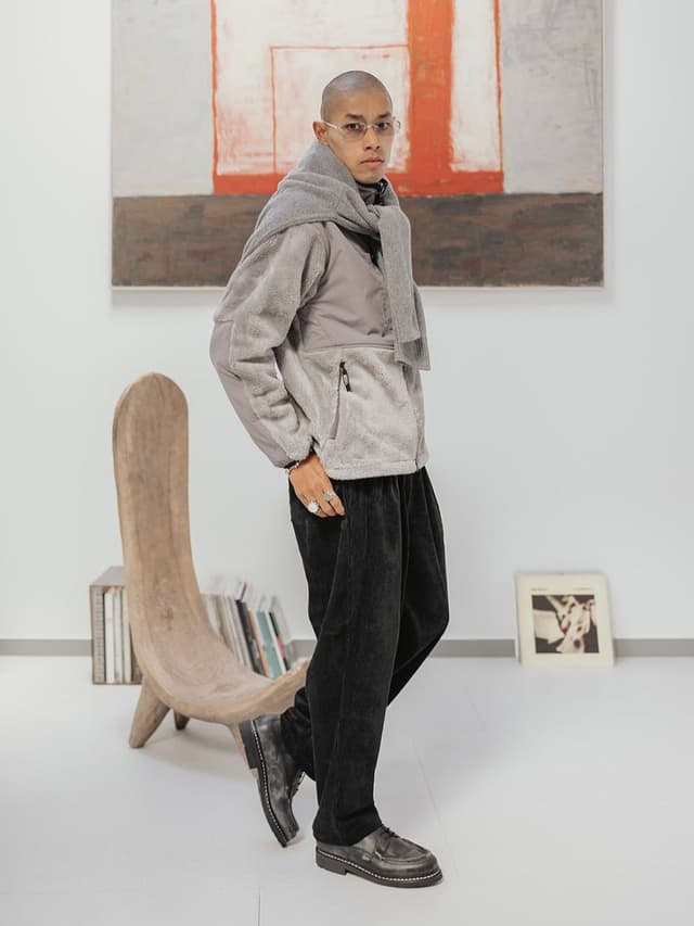 A man with a shaved head and glasses wears a grey fleece jacket, a grey scarf, black pants, and black shoes, standing in a minimalist room with an abstract painting.