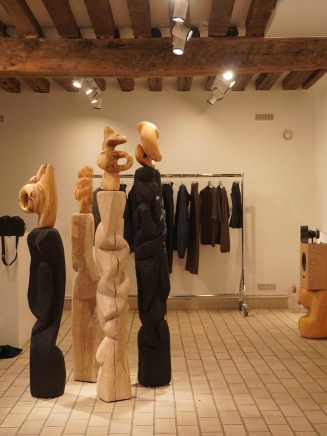 Four abstract wooden sculptures, two dark and two light, stand in a room with a clothing rack and exposed wooden ceiling beams.