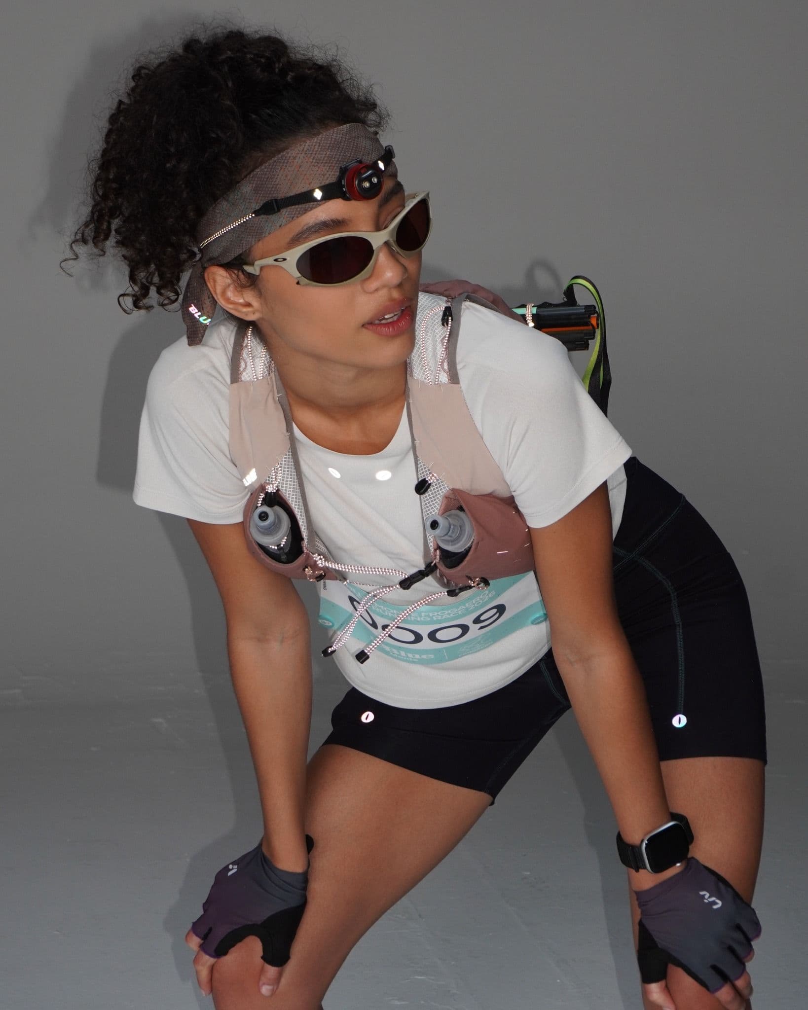 A female runner in a hydration vest, headlamp, sunglasses, and race bib leans forward with hands on her knees.