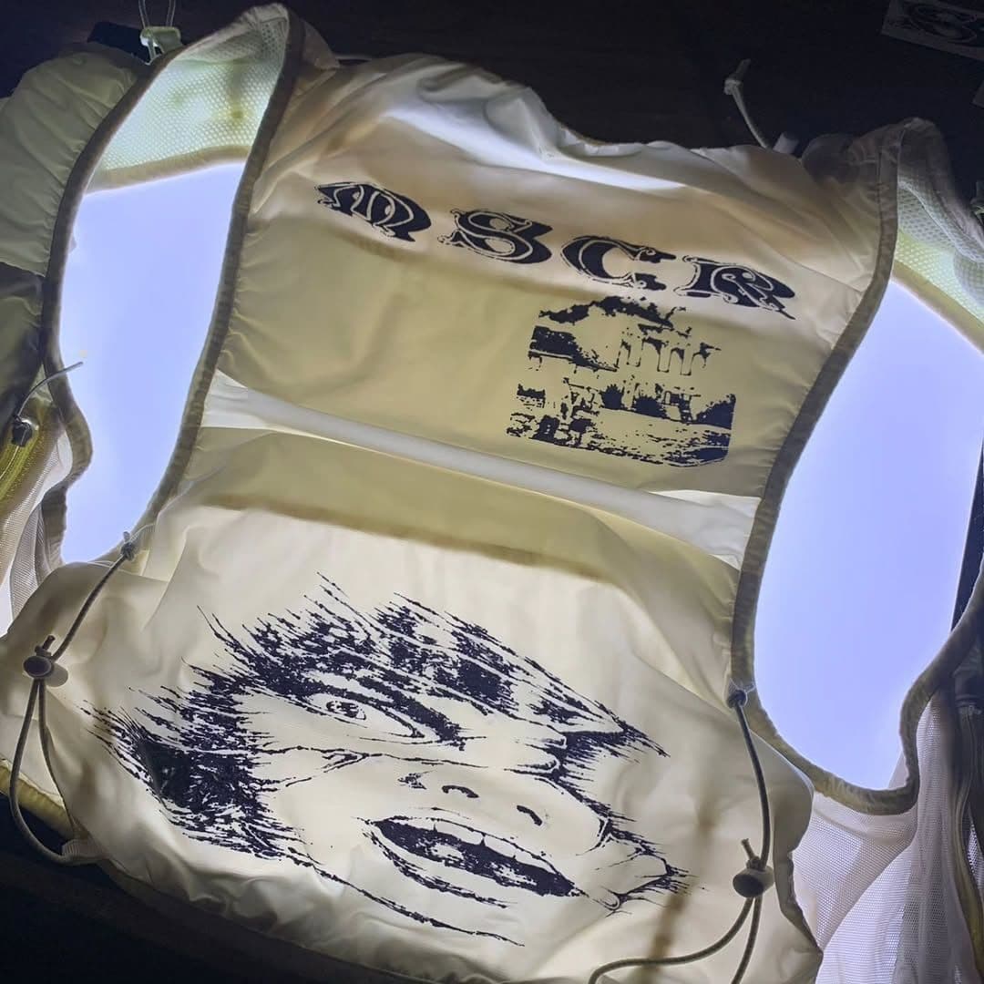 Backlit light-colored vest showing black graphics: "OSCR" text, an ancient building, and a stylized, unsettling face.