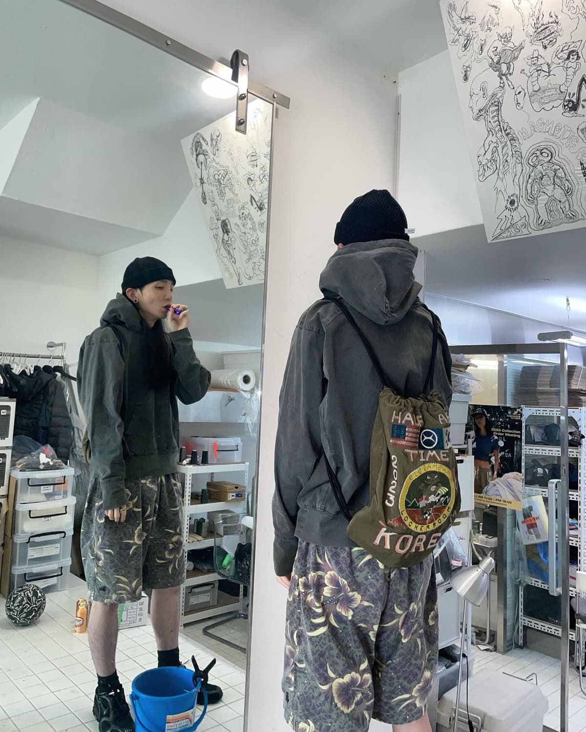 A person in a black beanie, grey hoodie, and floral shorts brushes their teeth in a mirror, carrying a green drawstring bag.
