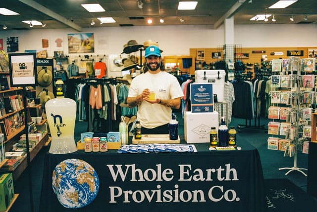 A smiling man in a blue hat stands behind a "Whole Earth Provision Co." display table with Nuun products, holding a drink in a retail store.