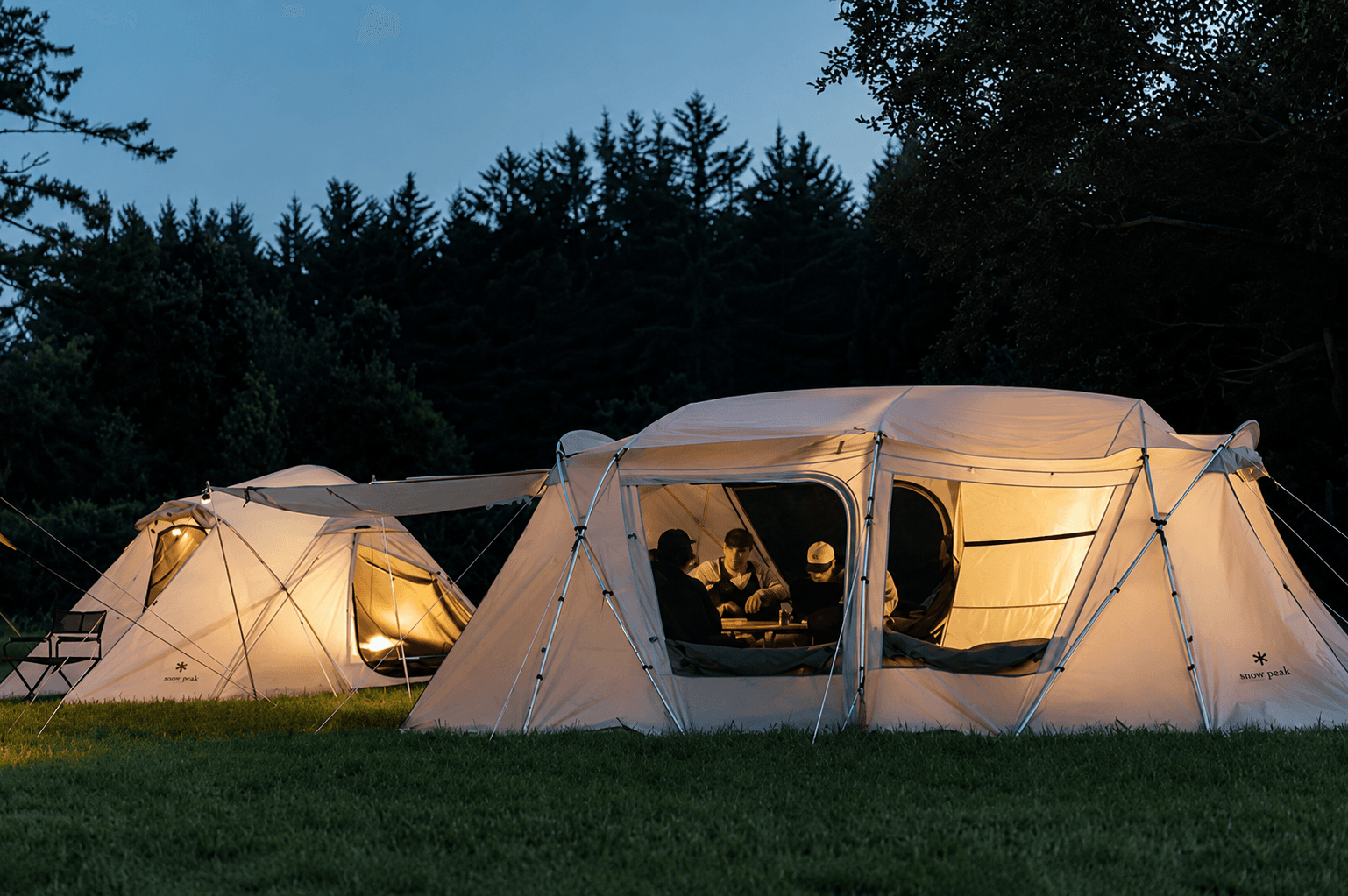 Two illuminated Snow Peak tents in a grassy field at dusk, with people inside the larger tent.