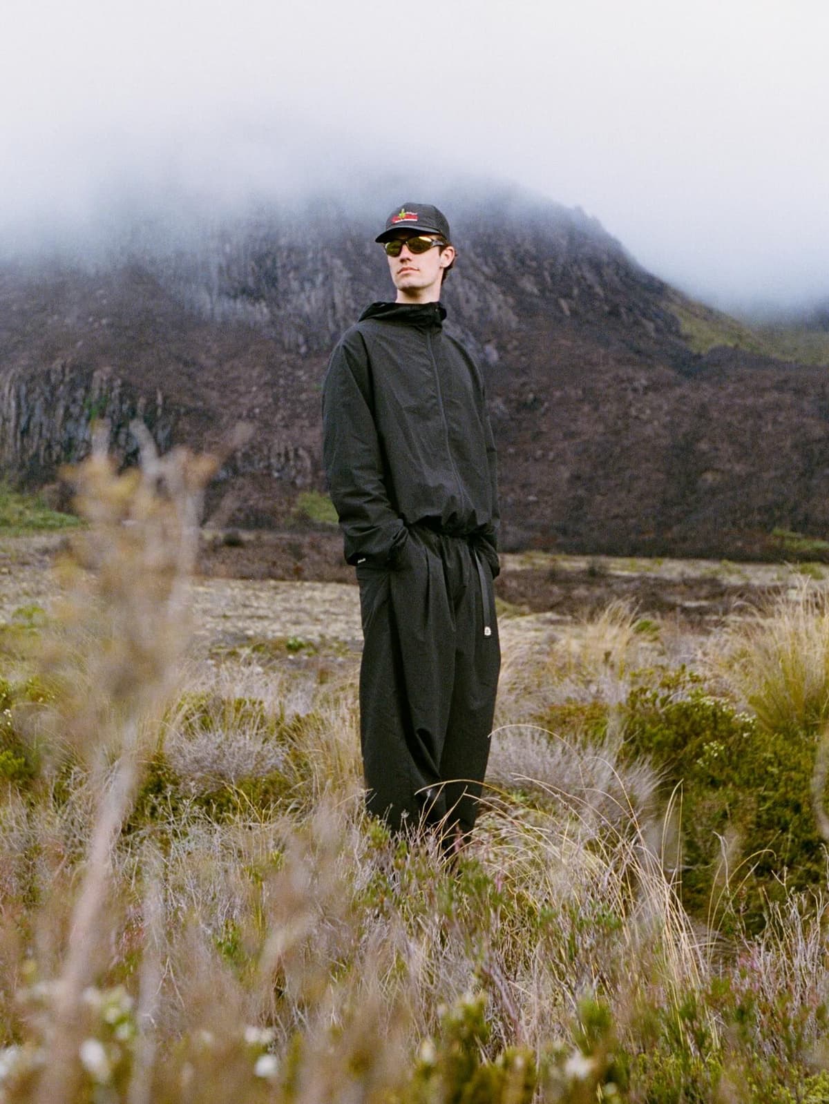 A man in a black tracksuit, cap, and sunglasses stands in a misty, mountainous landscape.