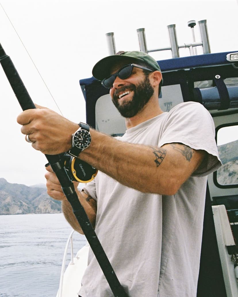 A smiling man on a fishing boat, holding a rod with mountains in the background.