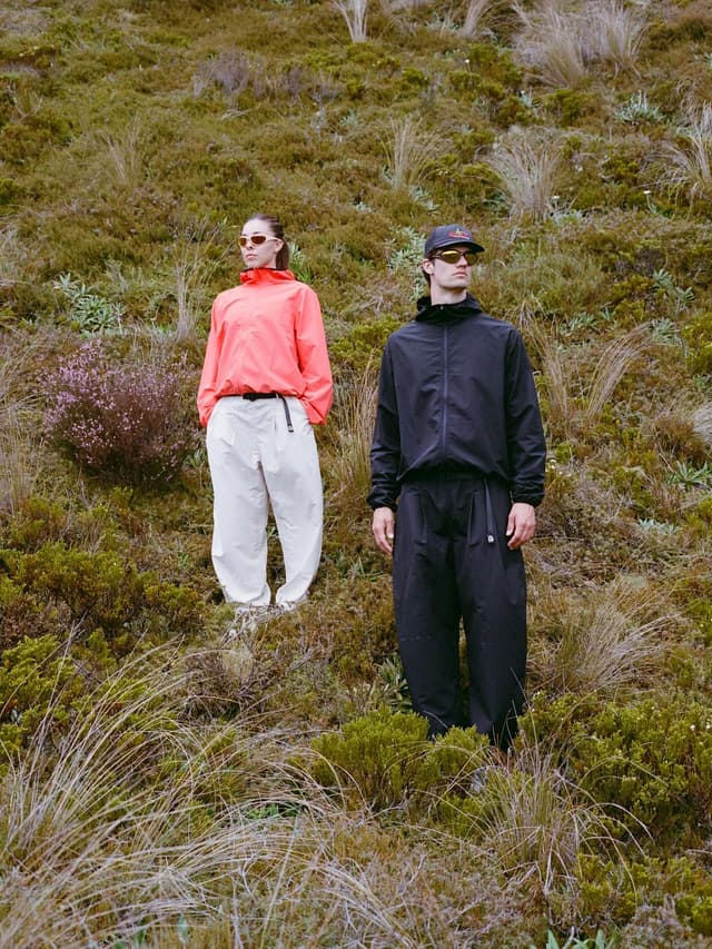 A woman in a coral jacket and white pants stands next to a man in a black jacket and pants, both wearing sunglasses, on a grassy hillside.