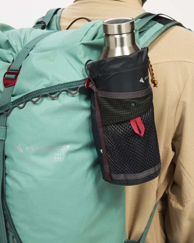 A person wearing a mint green backpack with a black water bottle holder attached, holding a silver water bottle.