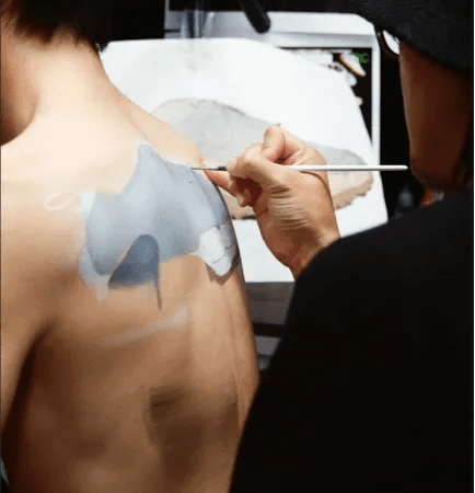 An artist paints a grey and white design on a person's shoulder with a fine brush.
