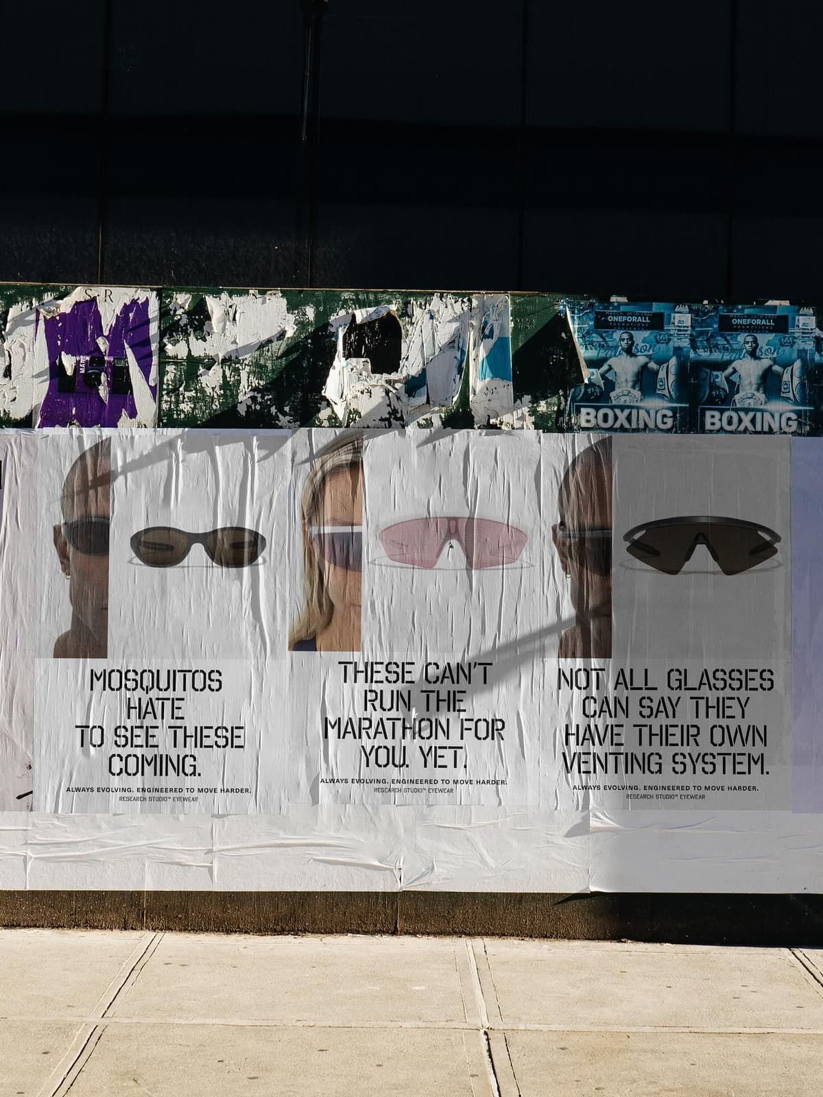 A large white poster on a street wall advertises sunglasses, featuring three faces wearing different styles and promotional text. Above it are torn posters; below, a concrete sidewalk.