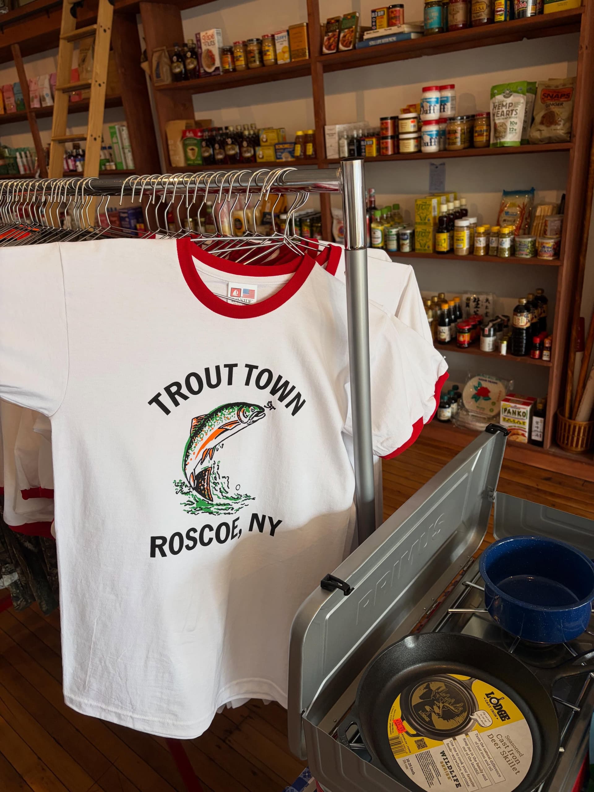 A white t-shirt with "TROUT TOWN ROSCOE, NY" and a jumping trout graphic hangs on a rack in a store, with shelves of food and a camping stove.