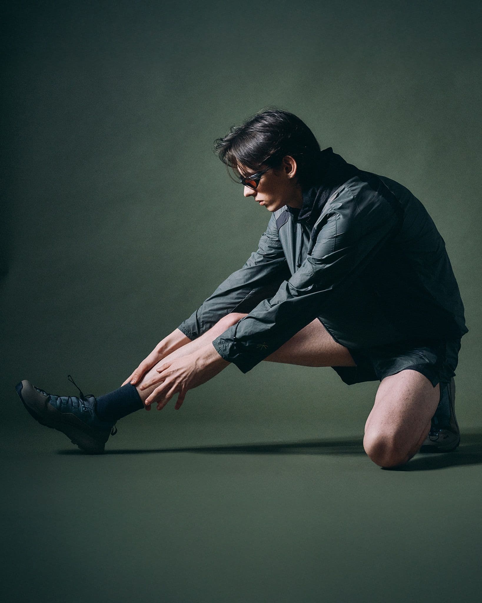 A person in dark activewear and sunglasses kneels and stretches, reaching their hands towards an extended foot.