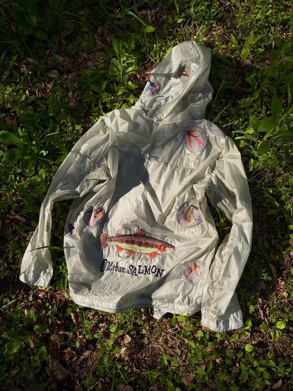Pale hooded jacket with embroidered salmon, fly fishing lures, and '@Urban.SALMON' text, on grass.