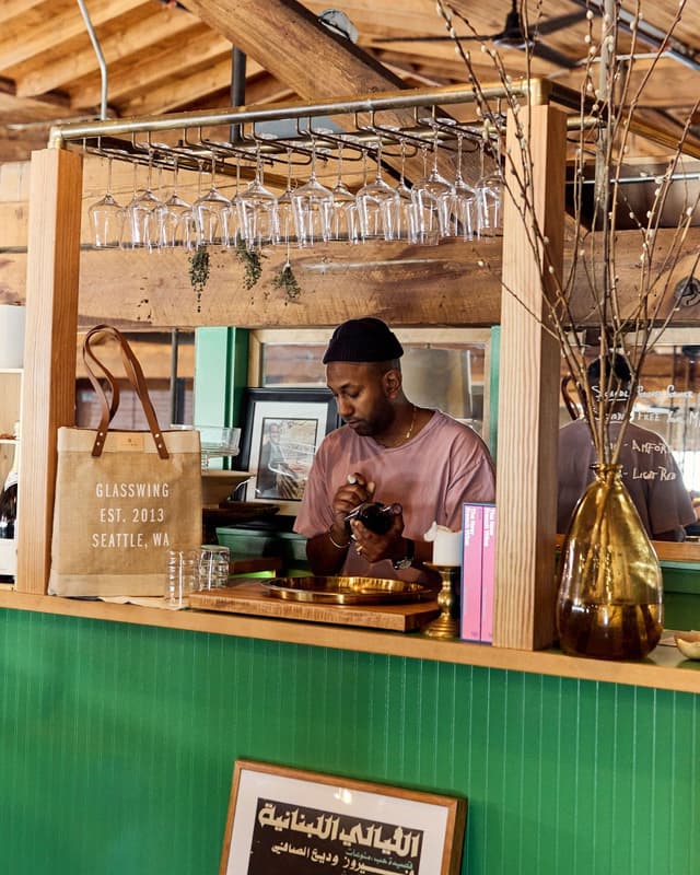 A man in a black beanie stands behind a green counter, holding an item, with wine glasses hanging overhead and a "Glasswing Seattle" tote bag beside him.