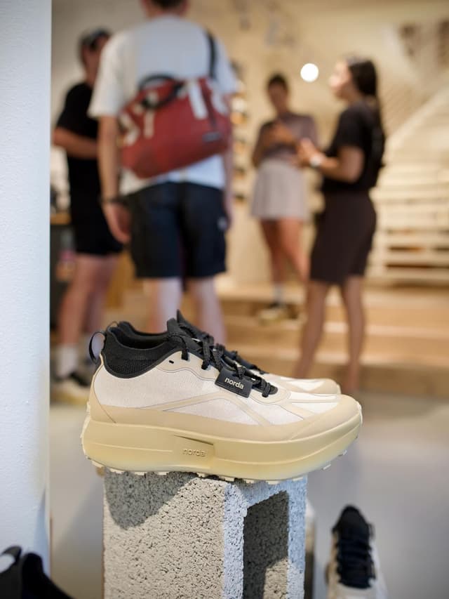 A pair of cream and black Norda running shoes displayed on a concrete block, with blurred people in a store background.
