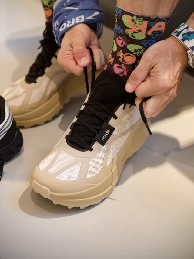 Hands tying the laces of a beige and white Norda running shoe, worn with colorful patterned socks.