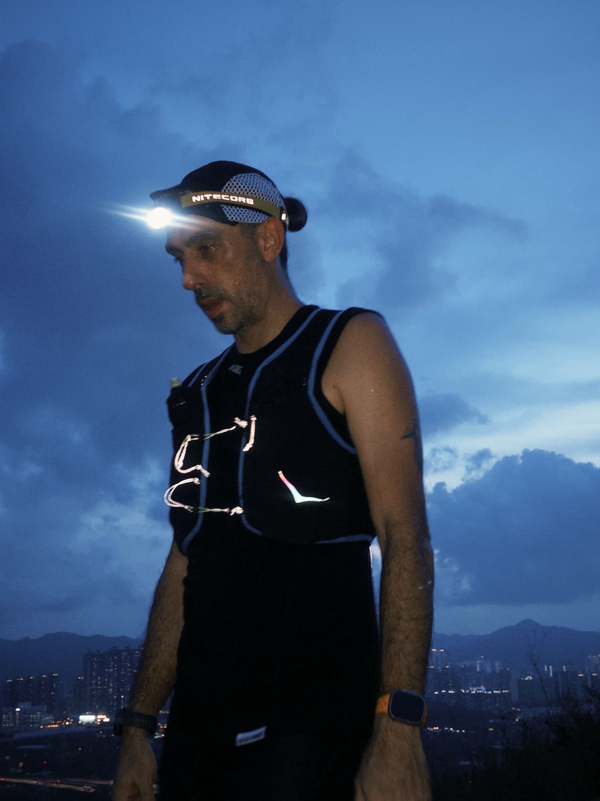 A man wearing a bright headlamp and reflective running vest at night, with city lights in the distance.