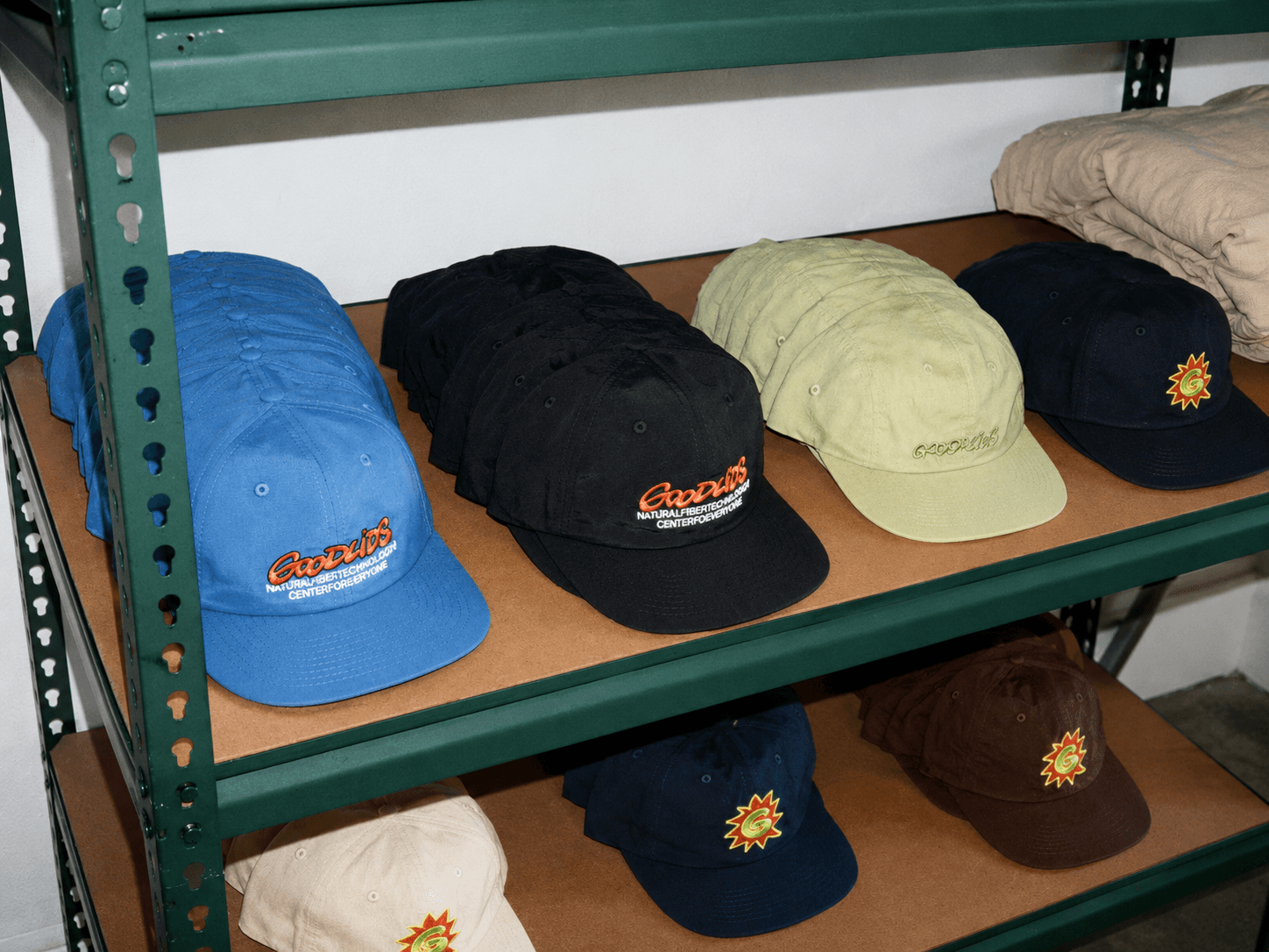 Rows of baseball caps in various colors, featuring "Goodwins" and sunburst logos, displayed on shelves.