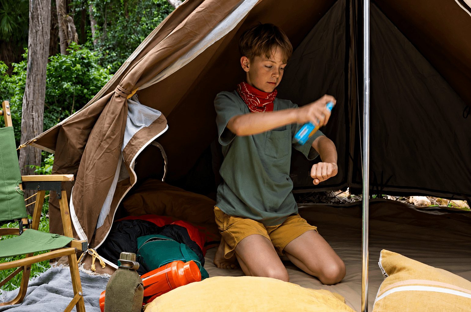 A boy kneeling inside a tent, spraying insect repellent on his arm.