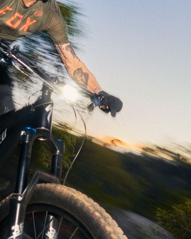 Close-up of a mountain biker's arm on handlebars with a bright headlight, motion-blurred background.