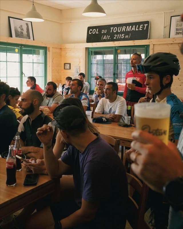 A group of cyclists relaxing in a rustic cafe at Col du Tourmalet, with the mountain pass sign visible on the wall.