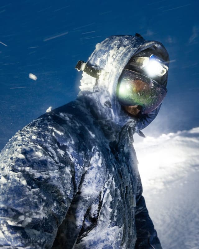 Person in a snow-covered jacket, glowing goggles, and headlamp in a blizzard.