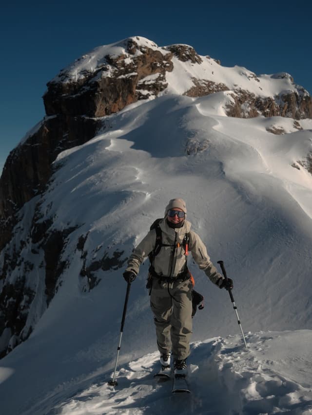 A person ski touring uphill on a snowy mountain ridge with a large snow-capped peak in the background.