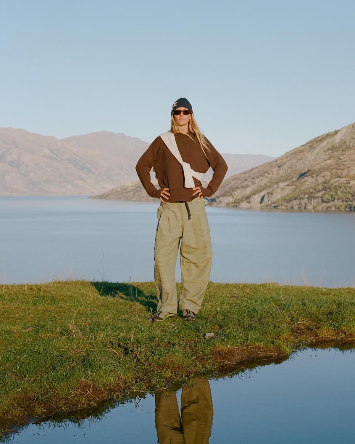 A woman in a brown sweatshirt, baggy pants, beanie, sunglasses, and a white sweater tied over her shoulders stands on a grassy bank beside a lake with mountains in the background.