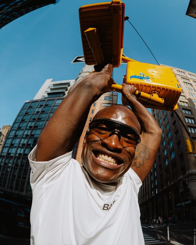 A grinning man in sunglasses holds up a yellow traffic light on a city street, captured with a fisheye lens.