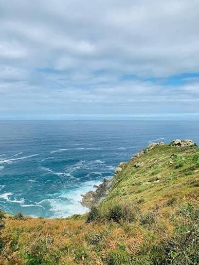 View from a cliff overlooking a vast blue ocean with white waves crashing against the rocky shore and a partly cloudy sky.
