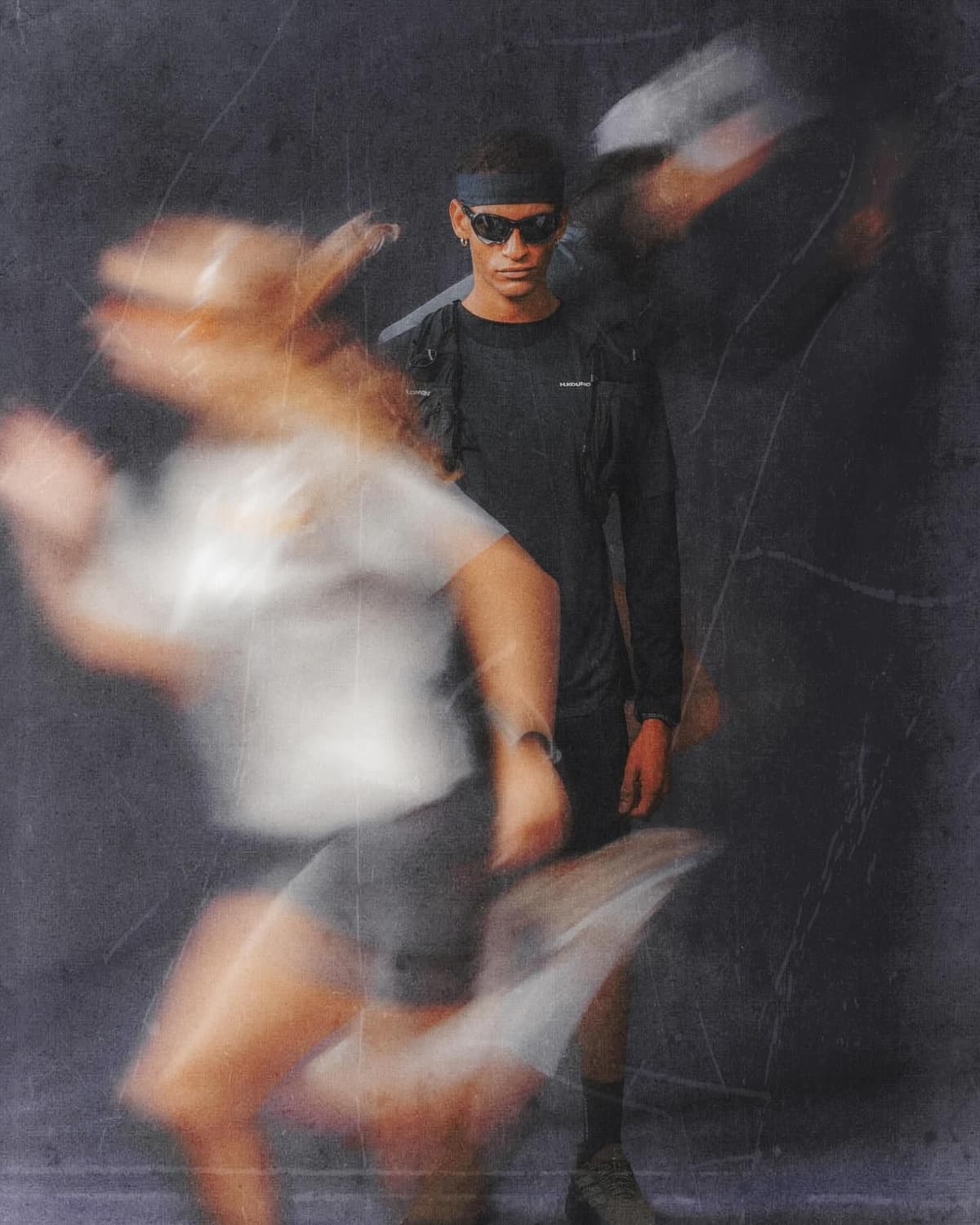 A static person in sunglasses and a headband stands behind a blurred figure in motion.