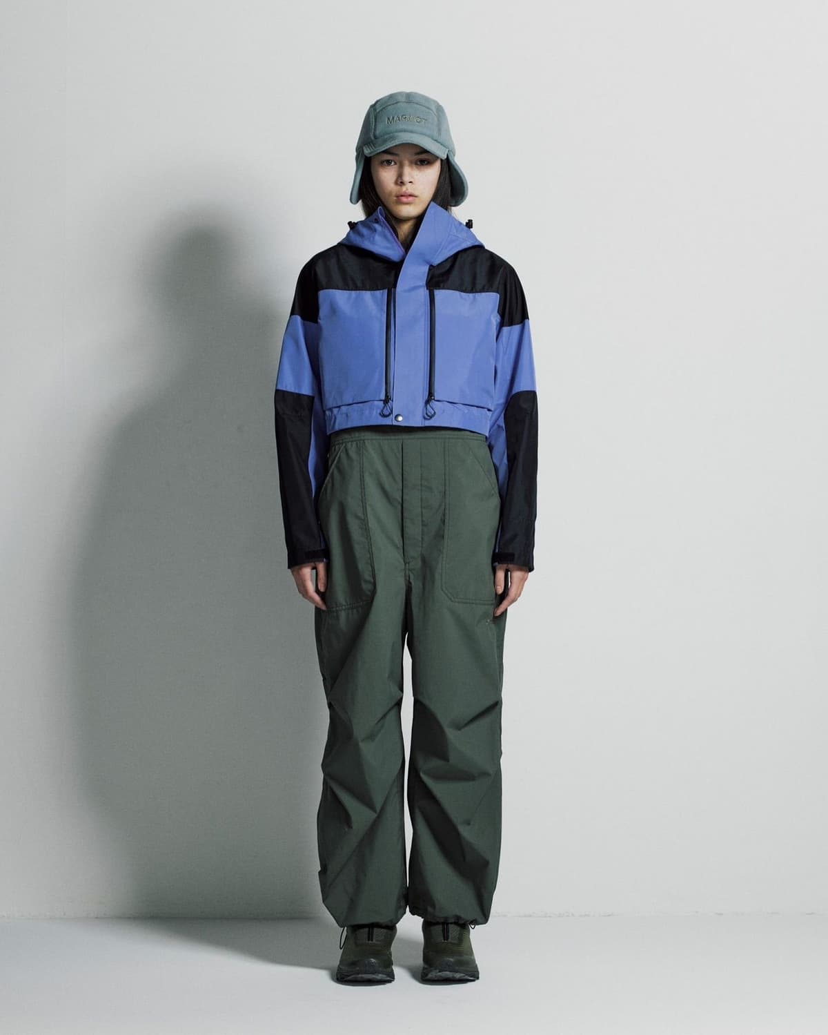 A woman wears a cropped blue and black color-block jacket, olive green pants, and a teal trapper hat.