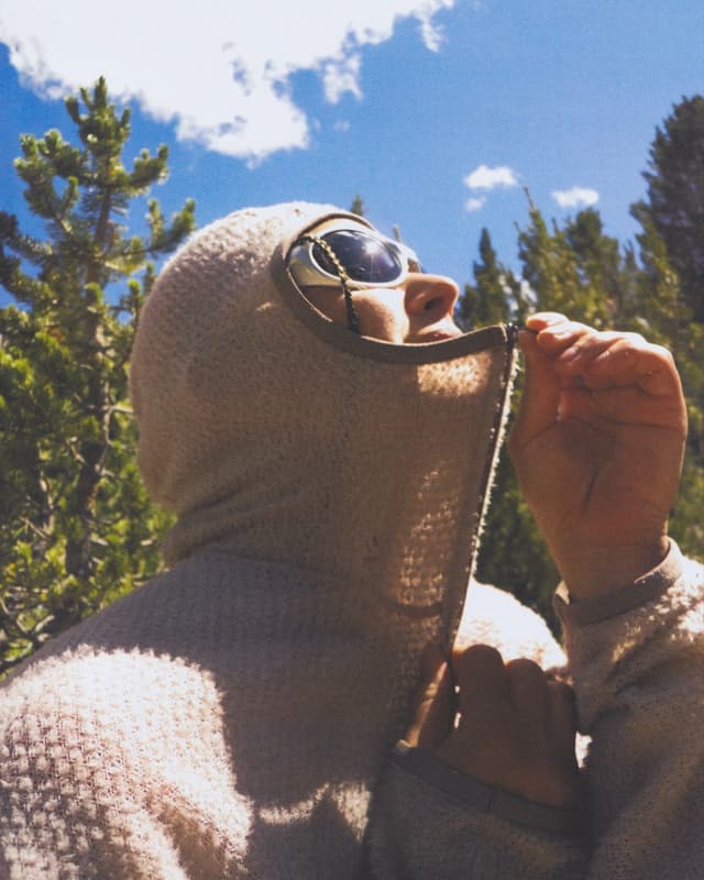A person in a light-colored, textured balaclava with round sunglasses looks up at a blue sky, pulling down the fabric at their chin. Trees are visible.