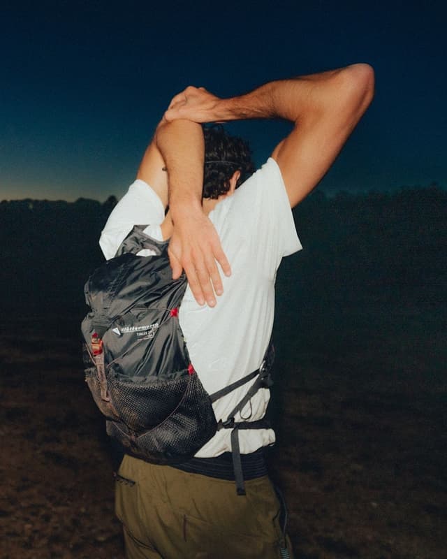 A person wearing a black backpack stretches their arms overhead against a dark sky.